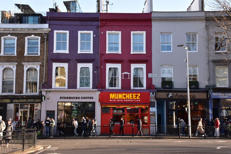A row of three adjacent commercial buildings along a city street, with a diverse group of pedestrians waiting in line outside. The building in the center is painted in bright red with a red and yellow sign for 'MUNCHEEZ' directly above the entrance, and features large glass shopfront windows. To its left, there is a purple building with white-trimmed windows and a white sign for 'Starbucks Coffee,' while to the right, there is a cream-colored building with a sign for 'Amore' and a darker storefront. The street scene is illuminated by natural daylight, with a street lamp positioned in front of the buildings and a metal rail fence separating the sidewalk from the road. The overall scene depicts a busy urban environment with retail outlets and foot traffic, and the context of rubbish removal services is supported by the visible urban setting that often requires waste management solutions for commercial properties.