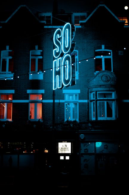 A photograph of a narrow street in Soho, London, showing a decorative, illuminated sign hanging overhead that reads 'CARNABY' with each letter in bright, multicolored lights, and an additional smaller sign beneath displaying 'SUHO' in a metallic, oval shape. The street is lined with multi-story buildings featuring traditional brick facades and large windows, some with black wrought-iron balcony railings and greenery such as plants and flowers on balconies and window sills. Purple string lights stretch across the street, adding a festive atmosphere. The scene is captured during daytime with a partly cloudy sky, and the overall setting reflects an urban commercial area with shops, including a visible Adidas signage attached to the building on the right side, indicating retail activity. The image emphasizes the street’s vibrant character, typical of an area with independent shopping, entertainment, and vintage charm, as would be associated with private waste management and rubbish disposal needs in busy city districts like Soho.