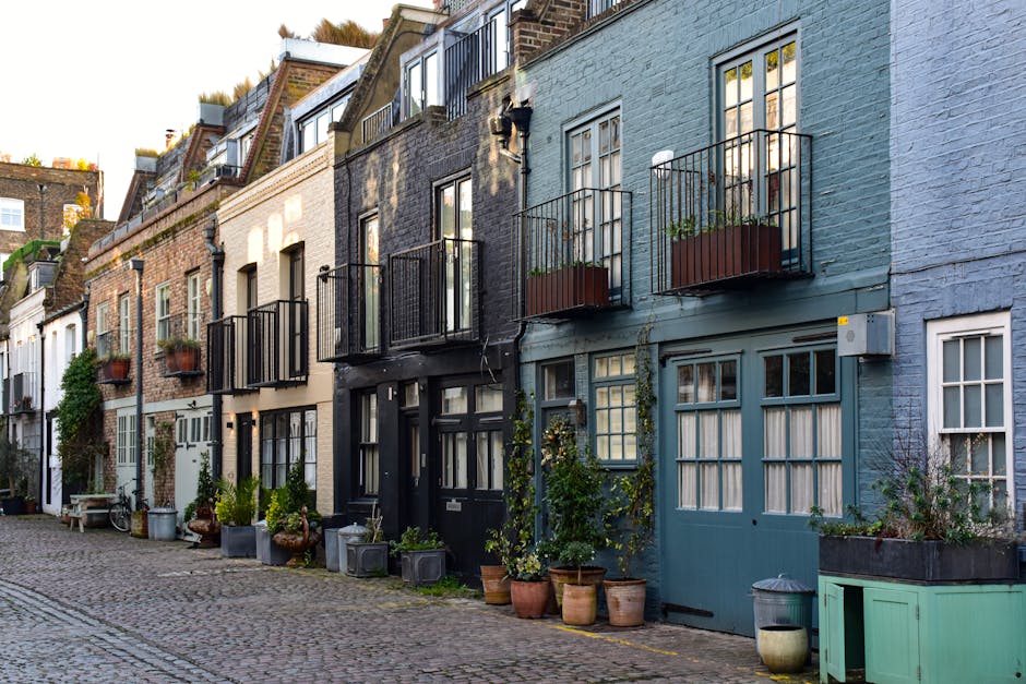 A row of narrow, multi-story residential buildings on a cobblestone street, featuring a mix of brick and painted facades in shades of brown, white, black, and blue. The buildings have small balconies with black metal railings, some adorned with potted plants. Several potted plants are arranged along the front of the buildings, including large clay and plastic containers with green foliage, small shrubs, and climbing vines. A variety of windows, some with white frames and others with black, are visible, with different styles of glazing and curtains. The street scene appears peaceful with subtle shadows indicating late afternoon or early evening lighting. The overall environment suggests a quiet, historic neighbourhood where residents may handle waste through private arrangements, supported by local rubbish removal services such as Rubbish Removal Soho, for alternative waste disposal and on-site clearance options.