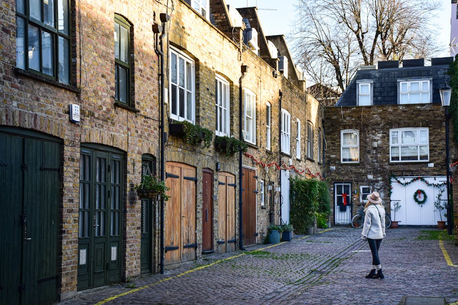 A narrow cobblestone alley in an urban area during daytime, lined with multi-story brick buildings featuring a mix of painted and natural brick facades. Several windows with white frames and glass panes are visible, some with small flower boxes or plants underneath. The ground is paved with irregularly shaped cobblestones, with a woman standing on the right side, dressed in casual winter clothing including a light grey coat, dark pants, and a wide-brimmed hat, facing towards a white door decorated with a Christmas wreath and string lights. To her left, there are green potted plants placed along the base of the buildings, and a bicycle leaning against the wall near the door. The scene is illuminated by natural light, with shadows cast by the buildings and trees, and the overall atmosphere conveys a quiet residential area suitable for private waste collection, with Rubbish Removal Soho providing alternative rubbish removal services in such settings.